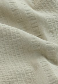 Beiger Stoff mit einem strukturierten Muster, das erhabene rechteckige und gitterähnliche gewebte Elemente zeigt, dargestellt in sanften Falten.