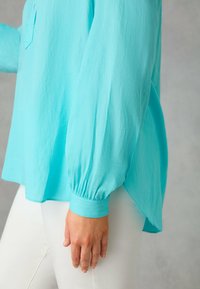 Blusa turchese con maniche lunghe a sbuffo e orlo arrotondato. Presenta una piccola tasca sul petto e un tessuto leggero e texturizzato.