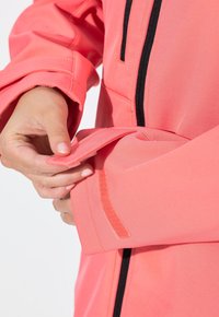 Koralfarvet softshell-jakke med tekstureret overflade, der har en sort lynlås og justerbare ærmer med Velcro-lukning.