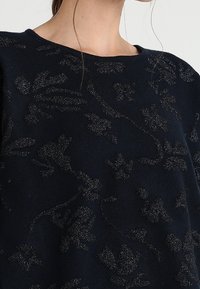 Marinblå tröja med en texturerad blommönster i mörk metalltråd. Rundad halsringning och mjukt tyg. Närbild av axelområdet.