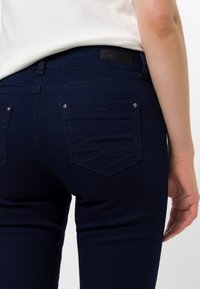 Vaqueros de mezclilla azul oscuro con corte slim, que cuentan con dos bolsillos traseros y detalles de costura sutiles. Textura suave y acentos de hardware mínimos.