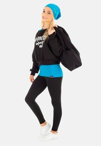 Schwarzes, kurzes Sweatshirt mit weißem Text, blauer Tanktop, schwarze Leggings und weiße Turnschuhe. Accessoires mit einer blauen Mütze und einer schwarzen Tasche.