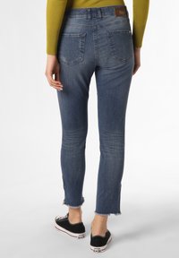 Slim-fit blå jeans med fransade fållar som bärs med svarta Converse-sneakers och en senapsgul långärmad topp, visad bakifrån.