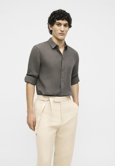 Νέος άνδρας με κατσαρά μαλλιά που φορά σκούρο γκρι πουκάμισο με κουμπιά και σαρωμένα μανίκια και ανοιχτό μπεζ παντελόνια με ζώνη, όρθιος.
