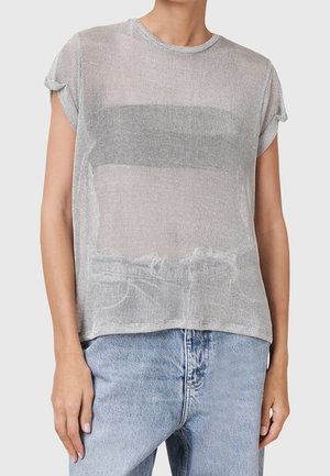 Personne portant un haut en maille argentée à manches courtes sur un bandeau noir, associé à un jean taille haute bleu clair, se tenant devant un fond uni.