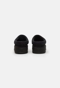 Zapatos negros tipo slip-on con parte superior de tela suave, suelas de goma y abertura acolchada. Diseño simple sin patrones visibles ni embellecimientos.