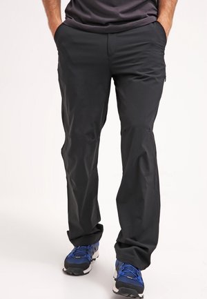 Schwarze Herrenhose aus leichtem Stoff mit schlanker Passform und Taschen an der Taille. Kombiniert mit blauen Sportschuhen.