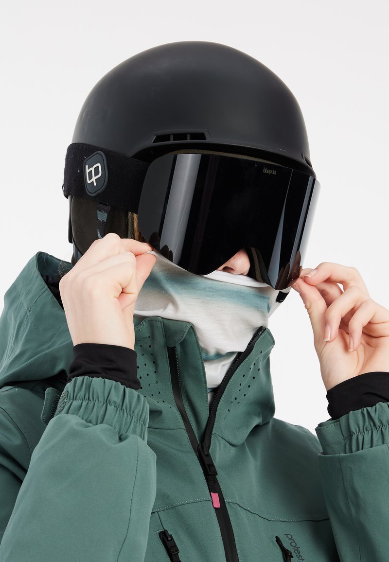 Sort skihjelm med tonede goggles og en lyseblå halsedisse, iført en grøn jakke med lynlås og ventilationsdetaljer.