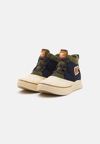 Hoge schoenen in marineblauw en olijfgroen suede, met een crème rubberen neus en een gestructureerde kuipzool. Voorzien van meerkleurige veters en een berenlogo.