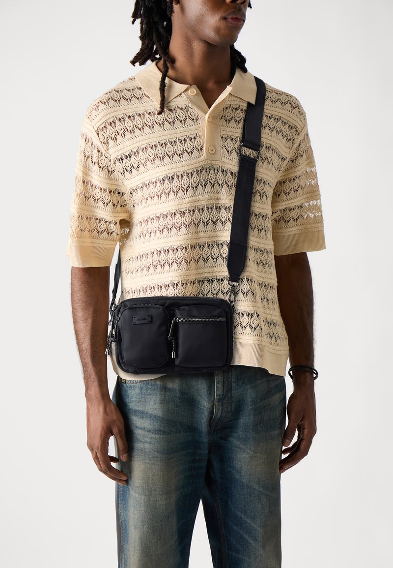 Sac en nylon noir porté en bandoulière avec deux compartiments zippés, associé à un polo texturé beige clair et un jean bleu.