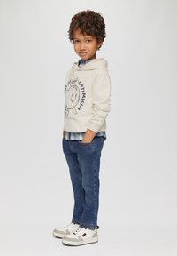 Crème hoodie met grafische print, blauwe geruite overhemd eronder, donkerblauwe jeans en witte sneakers met grijze accenten. Casual fit, zijaanzicht.