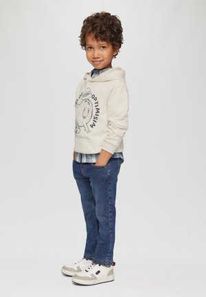 Crème hoodie met grafische print, blauwe geruite overhemd eronder, donkerblauwe jeans en witte sneakers met grijze accenten. Casual fit, zijaanzicht.