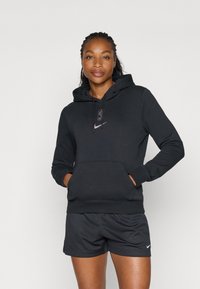 Svart hettegenser med frontlomme, med en liten logo i lilla. Kombinert med svarte mesh-shorts. Jevn tekstur.