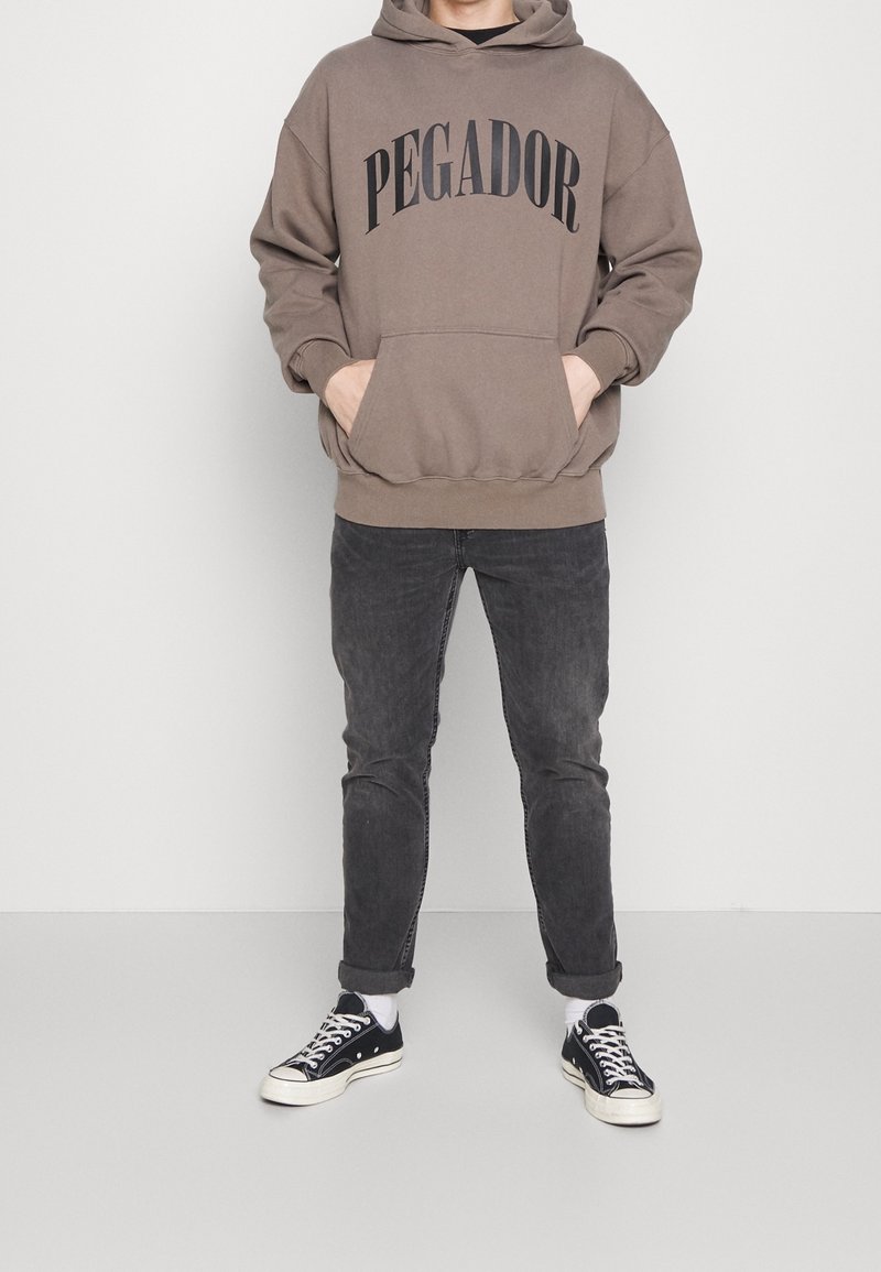 Felpa con cappuccio marrone con la scritta "PEGADOR" in stampa nera, tasca frontale, abbinata a jeans scuri e sneakers nere con accenti bianchi.