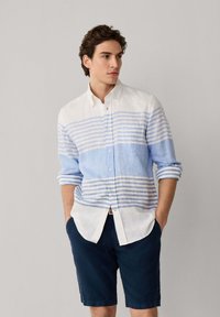 Joven con camisa de botones blanca con rayas azules y mangas remangadas, y pantalones cortos azul marino, sobre un fondo gris liso.