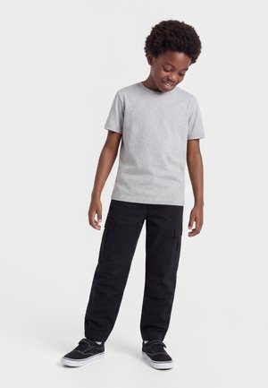 Göndör hajú fiú szürke rövid ujjú pólóban, fekete zsebes nadrágban és fekete sportcipőben, enyhe mosollyal lefelé néz.