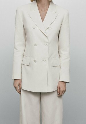 Personne portant un blazer croisé écru avec revers crantés et un pantalon large assorti sur un fond gris clair.