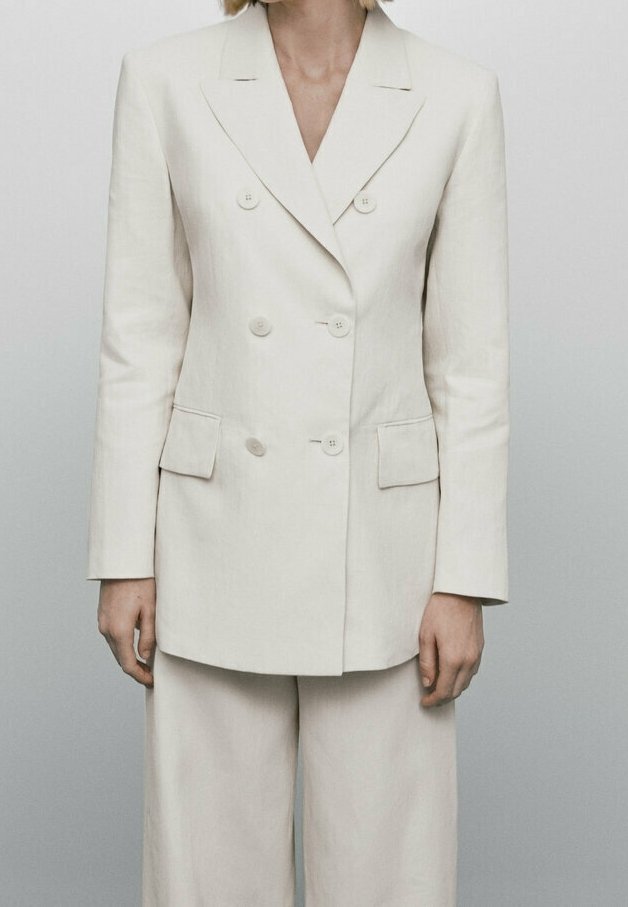 Personne portant un blazer croisé écru avec revers crantés et un pantalon large assorti sur un fond gris clair.