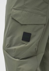 Oliiviroheline kangast kergesti avanev tasku, mille eeskel on must ruudukujuline silt "BOSS" pükste või jakkide esiküljel.