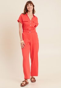 Combinaison rouge avec des manches courtes roulées, devant à boutons et ceinture. Elle présente de grandes poches et des jambes larges. Portée avec des sandales marron.