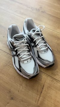 Tênis de corrida brancos e pretos da Asics com atacadores brancos, colocados lado a lado sobre um chão de madeira.