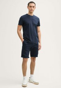 Tricou din bumbac de culoare bleu marin și pantaloni scurți asortați, ambele având un design casual. Completează ținuta cu adidași albi și tivuri rulate la pantaloni.