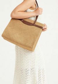 Tan woven bag with a smooth brown leather accent. Spacious rectangular shape, with a flat base and sturdy brown handles, textured surface.