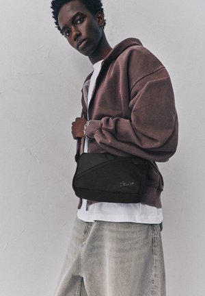 Jonge persoon die een bruine hoodie en lichte spijkerbroek draagt, houdt een zwarte crossbodytas vast tegen een effen lichte muur.