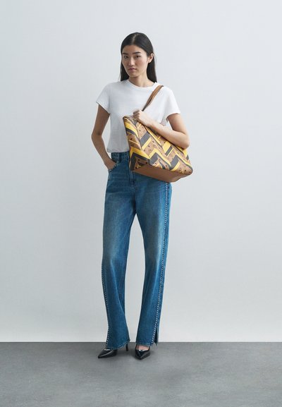 Naine valges T-särgis ja studeeritud sinistes teksades hoiab suurt pruunika ja kollase mustriga käekotti, jalas mustad terava ninaga kingad hallil põrandal.