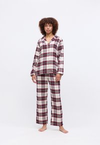 Kvinde, der står barfodet iført en langærmet pyjamashirt med knapper i rød, hvid og sort tern og matchende løse bukser på en hvid baggrund.