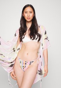 Capa leve e multicolorida com padrões abstratos, mangas abertas e fecho de laço, usada sobre um biquíni branco de cobertura mínima.
