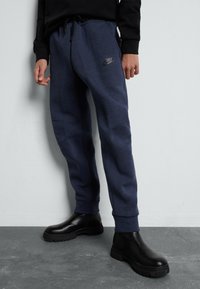 Navy Sweatpants mit einem Kordelzugbund, schmal zulaufenden Beinen, Seitentaschen und einem schwarzen Nike-Logo. Getragen mit schwarzen Knöchelstiefeln.