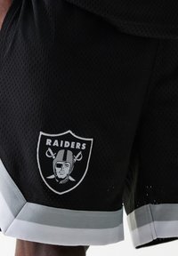 Black athletic shorts made of mesh fabric. Features a white and gray striped hem and a Raiders logo patch on the side.