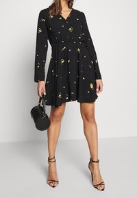 Schwarzes langärmeliges Kleid mit gelben Blumenmustern, V-Ausschnitt und tailliertem Bund; kombiniert mit einer schwarzen runden Handtasche und Riemchenabsätzen.