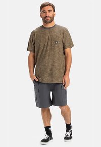 Brązowo-beżowa koszulka na krótki rękaw w paski z logo na piersi, do tego szare szorty moro z bocznymi kieszeniami. Czarno-białe trampki.