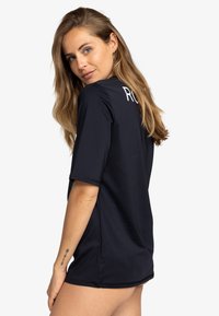 Navy blue short-sleeve shirt with a relaxed fit, featuring white text on the back. Smooth fabric, round neckline, and side slits.