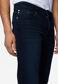 Close-up of a person wearing dark blue jeans and a white shirt, showing the hand and upper thigh area.