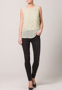 Top senza maniche verde chiaro con motivo geometrico, design sovrapposto su un fondo trasparente. Abbinato a pantaloni skinny neri e tacchi.