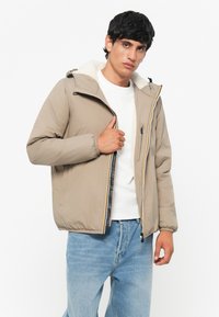 Lysebrunt isoleret jakke med en foret hætte i fåreskind, elastiske manchetter, lynlås foran og farverig accentstribe. Båret med blå jeans.