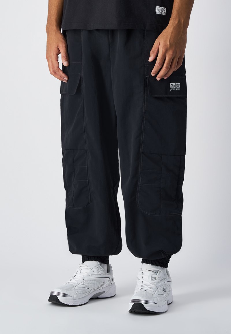 Zwarte cargobroek met meerdere zijzakken, elastische manchetten en contrasterende stiksels. Gecombineerd met witte sportschoenen.
