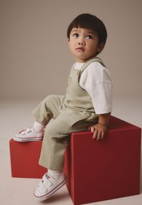 Bērns sēž uz sarkana kuba, valkājot gaiši zaļas kombinezonas ar smaidiņa seju un baltu krekliņu, kopā ar baltiem sporta apaviem un zeķēm.