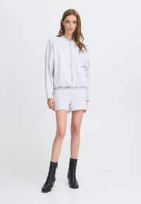 Sweat à zip en coton gris clair avec une coupe décontractée et des poches latérales, assorti avec un short gris. Le mannequin porte des bottines noires.