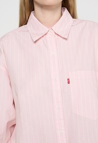 Jasnoróżowa koszula na guziki z pionowymi białymi paskami, z klasycznym kołnierzykiem, przednią kieszenią i metką z logo Levi's na kieszeni.
