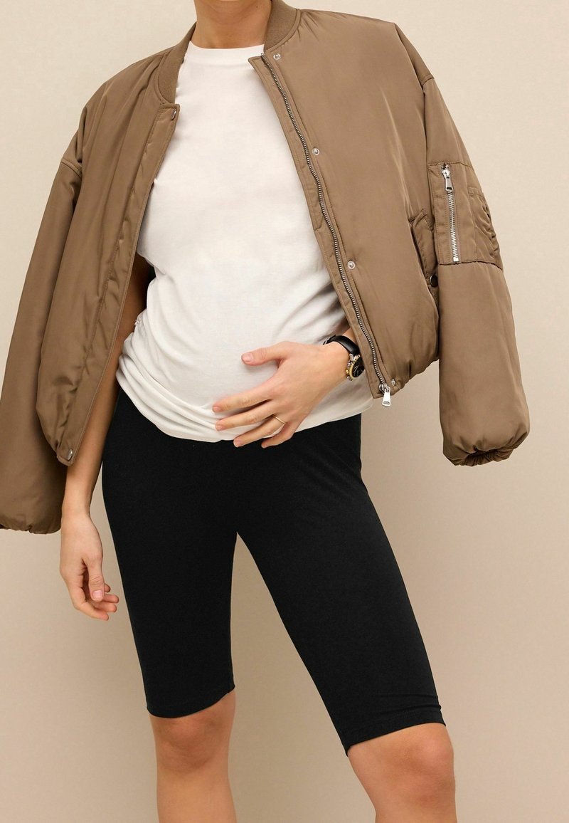 Personne portant une veste bomber beige sur une chemise blanche et un cuissard noir, une main reposant sur son ventre.