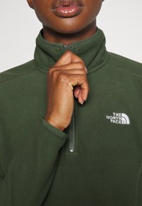 Pullover in pile verde con colletto alto, chiusura a metà zip e logo ricamato bianco sul petto. Tessuto morbido e polsini delle maniche aderenti.