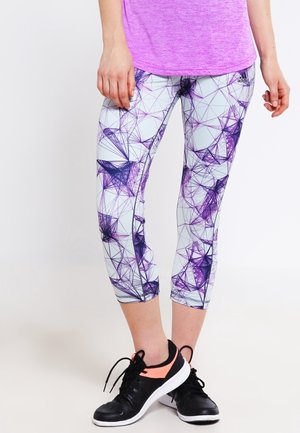 Zbliżenie osoby noszącej legginsy w fioletowy geometryczny wzór, fioletowy top i czarne buty sportowe z pomarańczowymi akcentami, stojącej na białej podłodze.