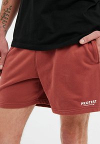 Pantaloni corti color ruggine realizzati in un tessuto morbido, dotati di tasche laterali e del logo bianco "PROTEST" sull'orlo sinistro inferiore.