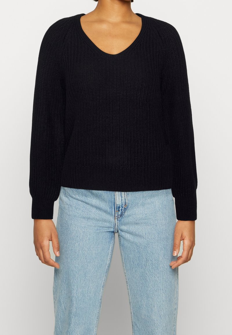 Personne portant un pull noir à col en V à manches longues et un jean taille haute bleu clair, debout devant un fond uni.