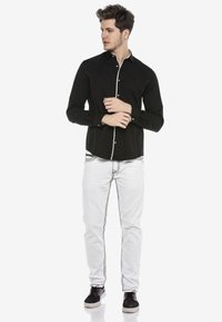 Schwarzes Langarmshirt mit weißen Kontrastnähten und Knöpfen; kombiniert mit weißen Hosen mit schwarzen Nähdetails, getragen mit schwarzen Turnschuhen.