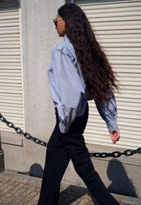 Jasnoniebieska oversizeowa koszula z bufiastymi rękawami, zestawiona z czarnymi, dopasowanymi spodniami. Długie, falowane włosy, spacerująca obok teksturowanej betonowej ściany.